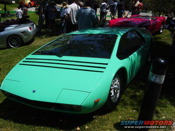 Teal sports car with pop-up headlights and black stripes on the hood, parked on grass