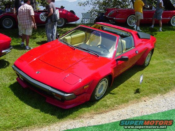 Red sports car with pop-up headlights and beige interior parked on grass at an outdoor event. Other cars and people are visible in the background