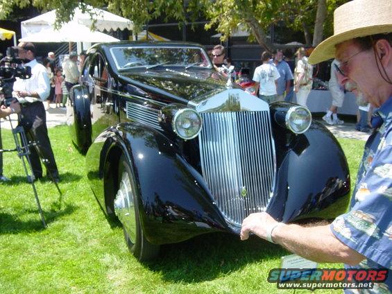 man in straw hat looking at a vintage black car with a tall chrome grille and round headlights