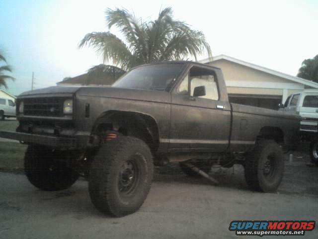 Lifted pickup truck with aftermarket bumper and large tires. Another white vehicle is parked behind it