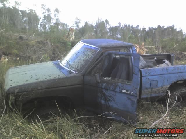 Muddy blue pickup truck with door open, stuck in tall grass and brush