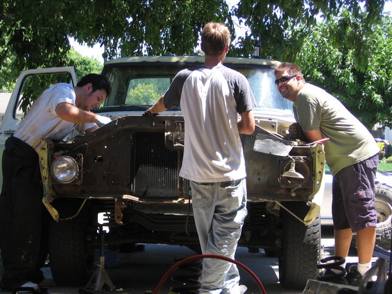 three guys working on the front end of a rusty truck, hood removed