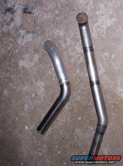 Two bent metal exhaust pipes on a concrete floor. One pipe is polished, the other has visible welds