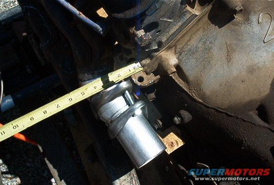 Engine block with a metal exhaust pipe and a yellow measuring tape. The tape shows approximately 2 inches