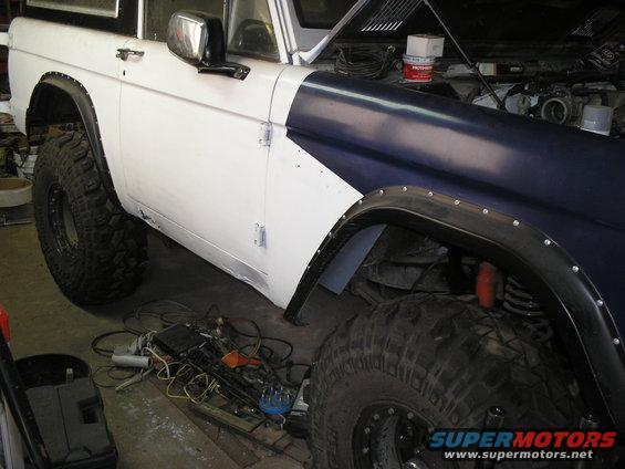 White and blue Ford Bronco with large tires in a garage, hood up, showing engine parts and tools on the floor