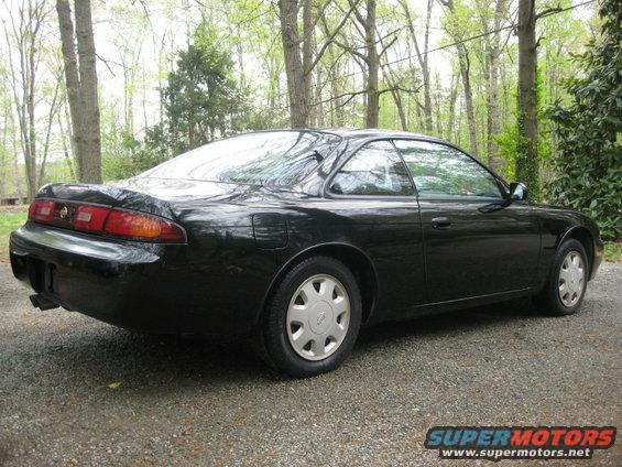 Black 1995 Nissan 240SX coupe from the rear-right quarter, parked on a gravel surface with trees in the background