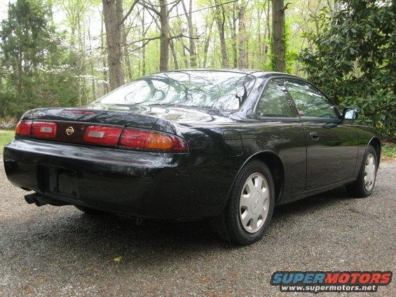 Black 1995 Nissan 240SX coupe, rear 3/4 view. The car is parked on a gravel surface with trees in the background