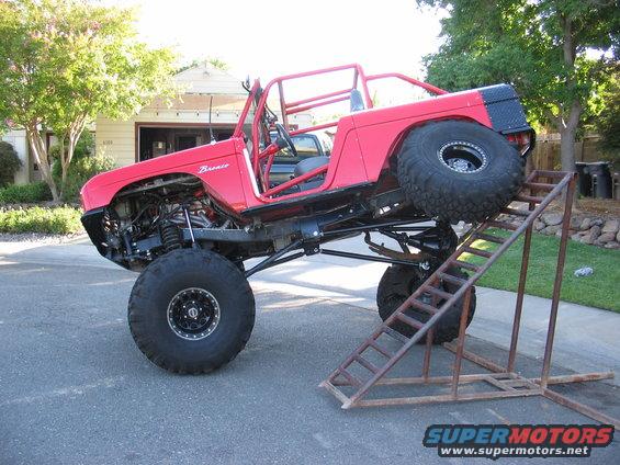 Red 1970 Ford Bronco with custom roll cage and large tires, climbing a metal ramp