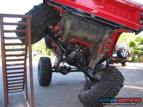 Red 1970 Ford Bronco chassis with large tires on a ramp, showing undercarriage and suspension