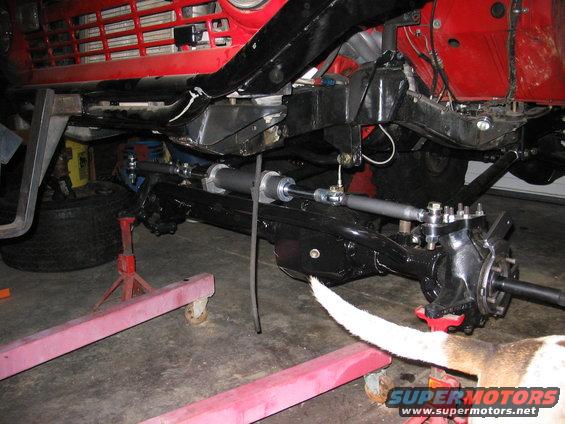 close-up of a vehicle's front axle, steering linkage, and suspension parts, with a red grille visible above