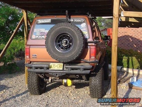 Rear view of a 1986 Ford Bronco II parked under a wooden shelter. It has a spare tire mounted on the back and a custom bumper