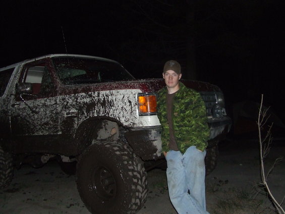 1990 Ford Bronco covered in mud, with a man in a camouflage jacket standing beside it at night