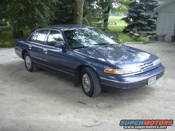 Blue sedan parked on dirt, front quarter view, chrome wheels, visible license plate
