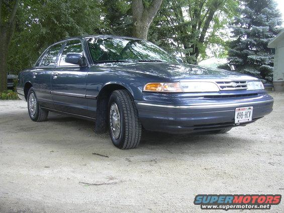 Dark blue Ford Crown Victoria parked on a dirt lot with trees and a house in the background. License plate reads 498-HCV
