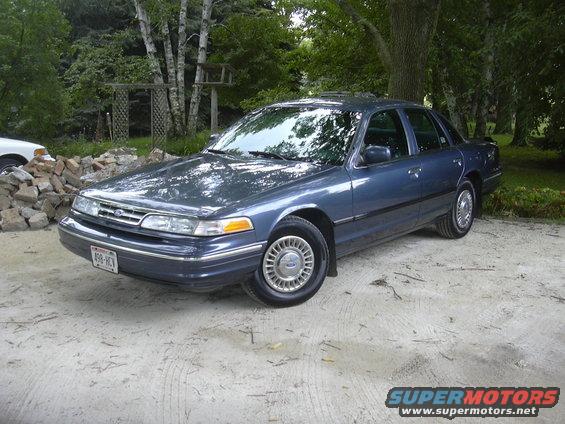 Blue sedan parked on dirt lot, trees and fence in background. License plate is visible