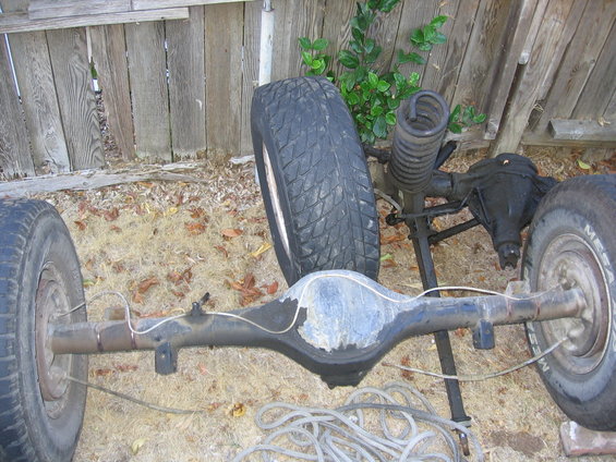 Vehicle axles and tires are laid out on the ground in front of a wooden fence