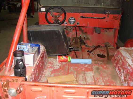 Interior of a red Jeep CJ with a black steering wheel and seats, floor is dirty