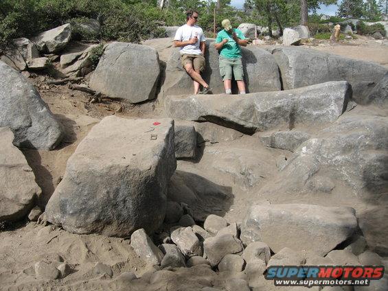 Two men stand on large rocks in a rocky outdoor area. Red and black markings on a rock