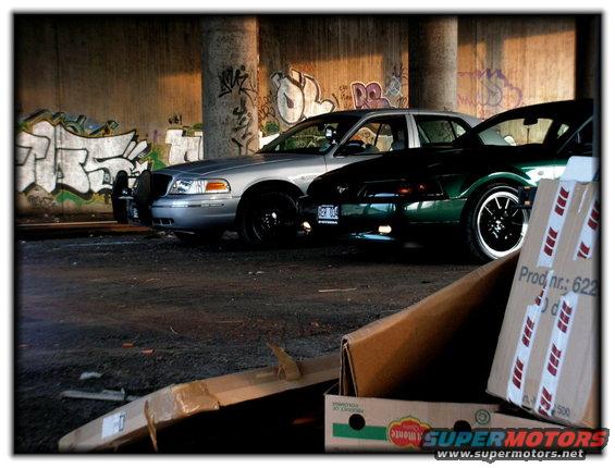 1998 Ford Crown Victoria and a green Ford Mustang parked under a bridge with graffiti