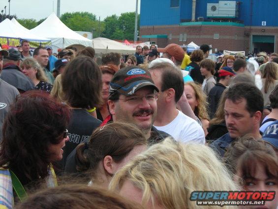 Crowd of people at an outdoor event, some wearing hats and t-shirts. Building in background