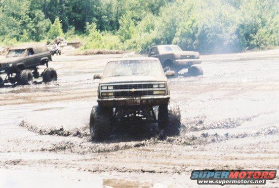 Lifted pickup truck with large tires driving through mud, another truck in background