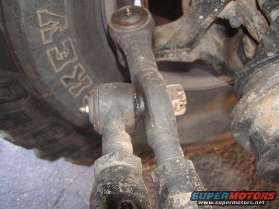 Close-up of vehicle steering linkage with tire in background. Text on tire reads 'KEW'