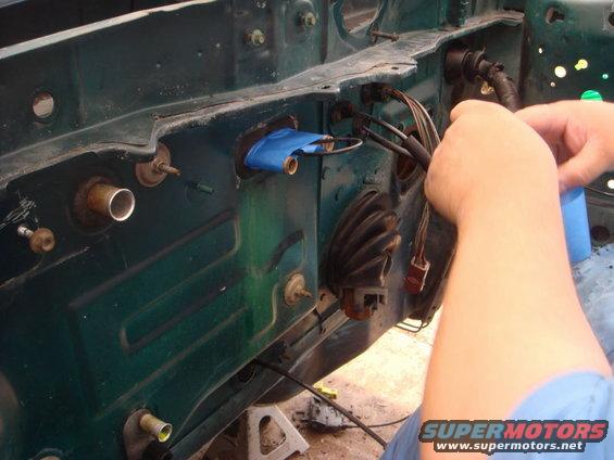 Car firewall with green paint, showing wiring harness and blue tape on a grommet. A person's hands are visible working on the wires