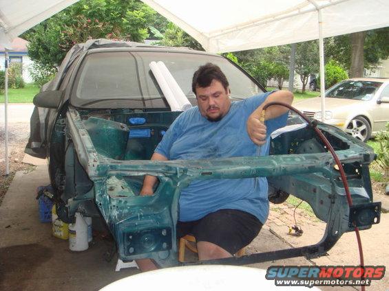 Man sits inside disassembled car frame, working with an air hose. Another car is parked behind