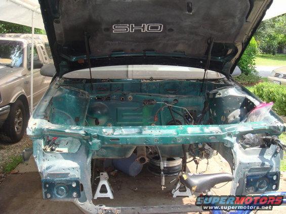 Green car engine bay with hood open, disassembled. SHO emblem visible on hood liner. Another vehicle in background