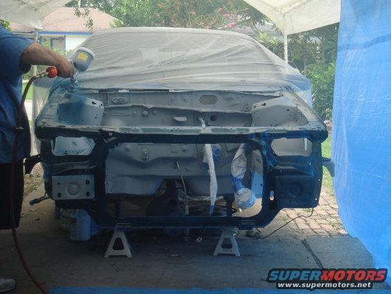 Car body on jack stands, being spray painted under a tarp. Front end is exposed