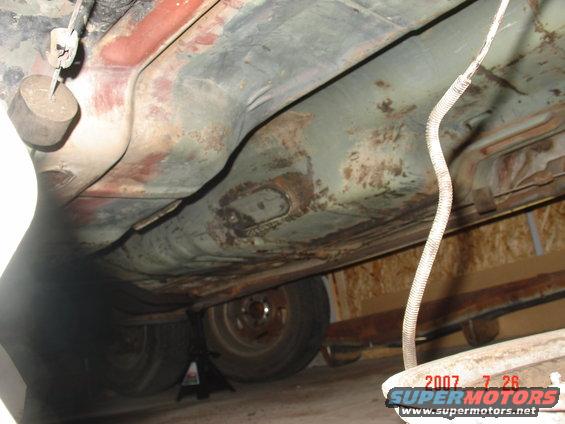 Underside of a vehicle showing rusty floorboards and some suspension parts. A wheel is visible on a jack stand