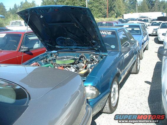 Teal 1987 Ford Mustang with hood open, showing engine. Parked next to a grey car and other vehicles in lot