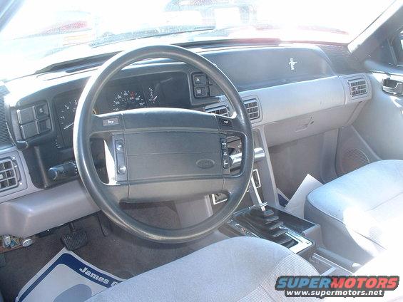 car interior with steering wheel, dashboard, and seats. The dashboard has gauges and vents. A floor mat is visible on the driver's side