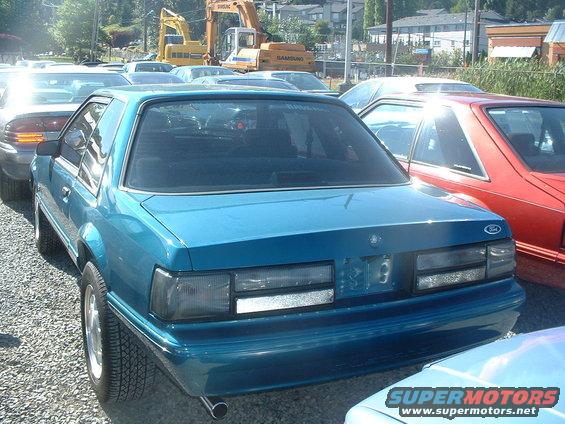 Blue coupe, rear view, showing taillights and Ford emblem. Other vehicles and construction equipment in background