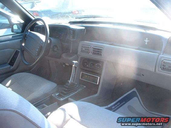 Interior of a car with a manual transmission, dashboard, and steering wheel. A Mustang emblem is visible on the dash
