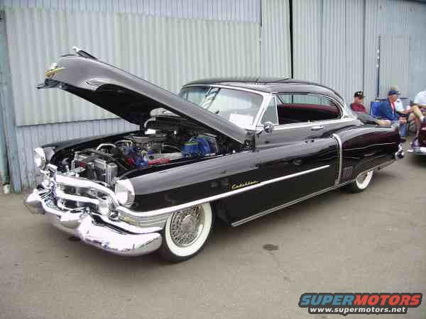 Black coupe with hood open, showing engine and chrome grille. Whitewall tires