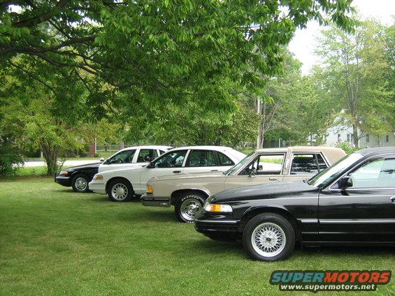 Four sedans parked in a row on a grassy lawn under trees. One is black, two are white, and one is tan