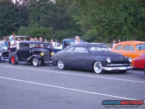 Several classic cars parked in a lot, including a black hot rod and a dark gray sedan with white wall tires