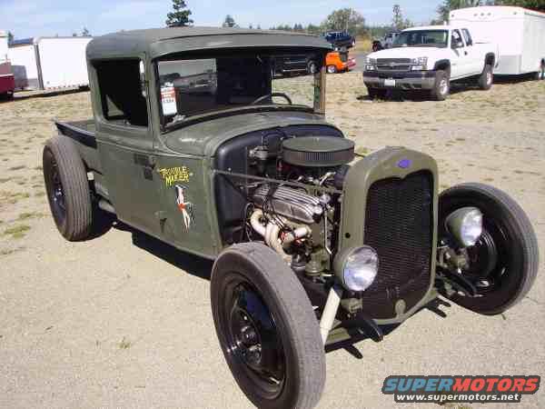 Olive green hot rod pickup truck with exposed engine and wide rear tires. Text on door reads 'Trouble Maker'