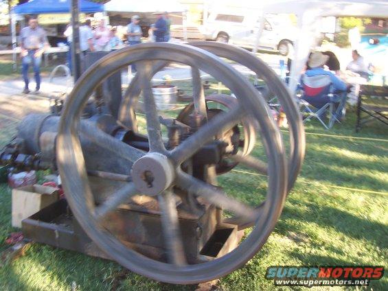 Close-up of an old engine with two large flywheels, set up outdoors with people and tents in the background