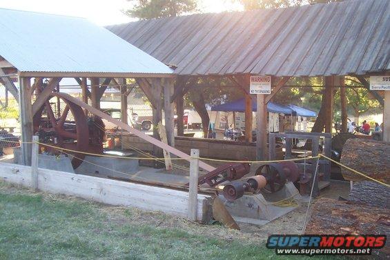 Outdoor scene with old industrial machinery under a wooden roof, logs nearby