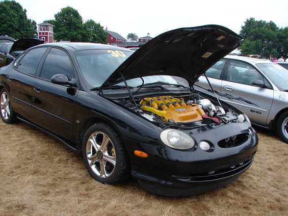 Black Ford Taurus with hood open, showing a yellow engine cover. Chrome wheels and tinted windows are visible