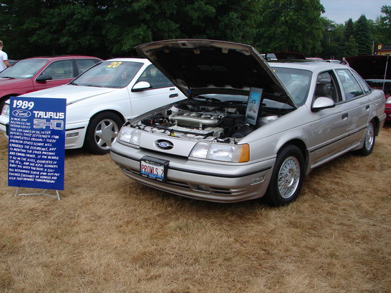 1989 Ford Taurus SHO with hood open, parked next to another car. Sign details the car's history