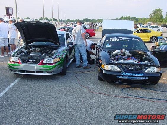 Two Ford Mustangs, one silver with green and red flames, one black with racing decals, hoods open at a drag strip