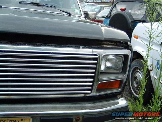 Front grille and headlight of a dark-colored Ford truck, parked next to a white vehicle