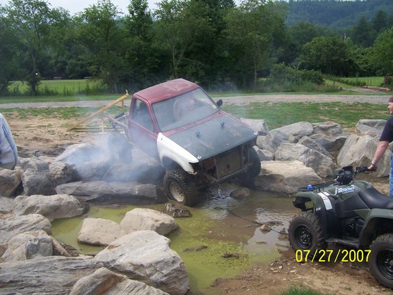 Red and white pickup truck with missing grille stuck in mud and rocks, an ATV is nearby
