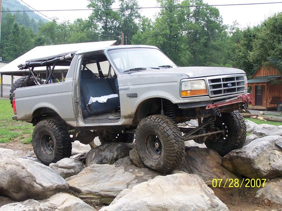Modified Ford Bronco with off-road tires navigating large rocks. The driver's door is open, revealing the interior