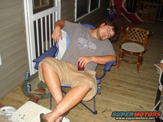 Man sleeping in a blue folding chair on a wooden deck, holding a red cup