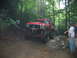 1992 Ford Mustang - A heavily modified red truck navigates a challenging muddy terrain in a wooded area.