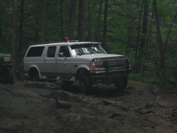 White Ford Bronco SUV on a rocky, muddy trail in the woods. Another green vehicle is partially visible to the left
